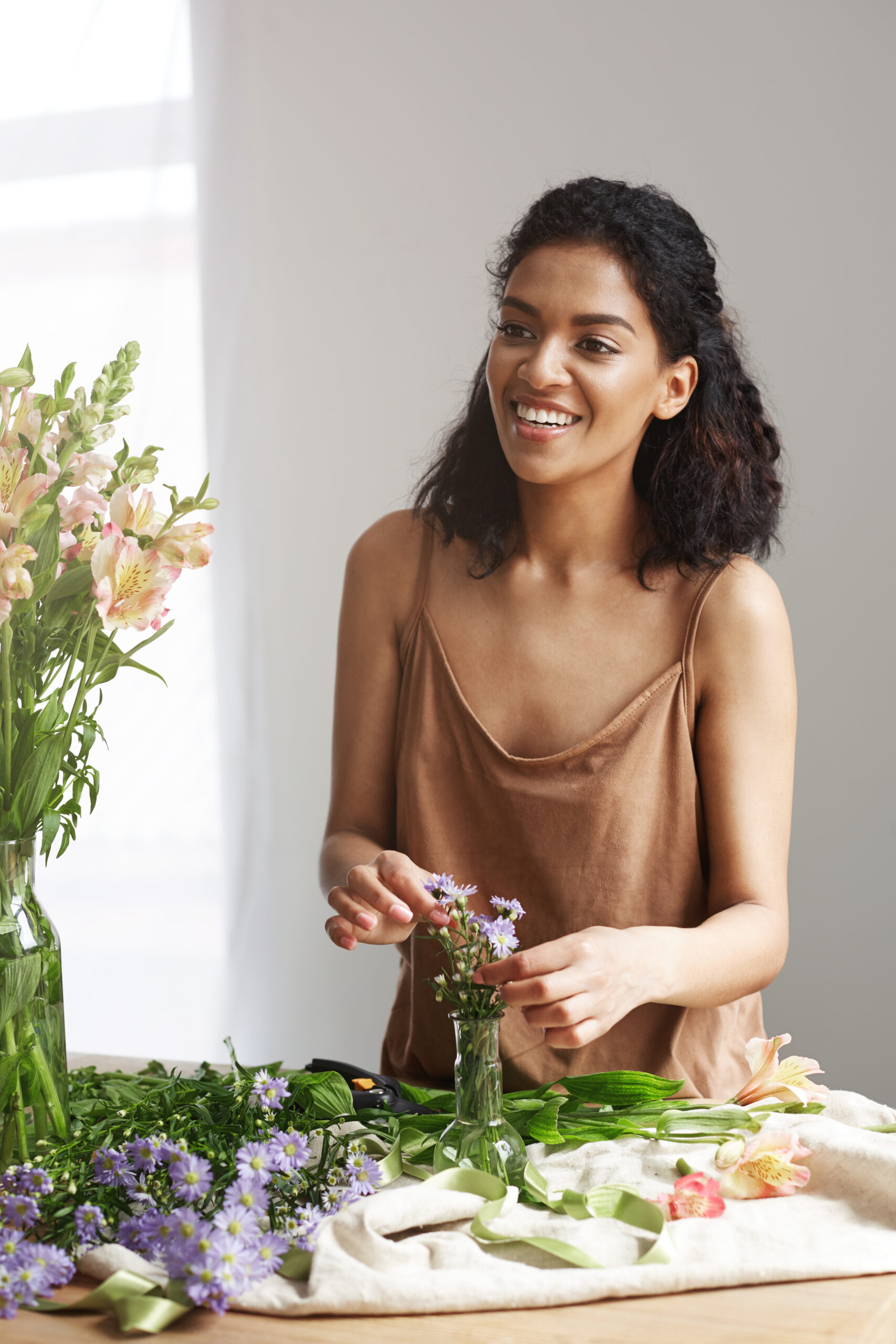 Young beautiful african girl florist smiling making bouquet in flower shop over white wall.